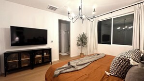 A modern bedroom with a large flat-screen TV, a wooden cabinet, a bed with a patterned pillow, and a potted plant. - New! 4BR Vacation Home, Tub, BBQ, EV, close to Sequoia (Visalia)