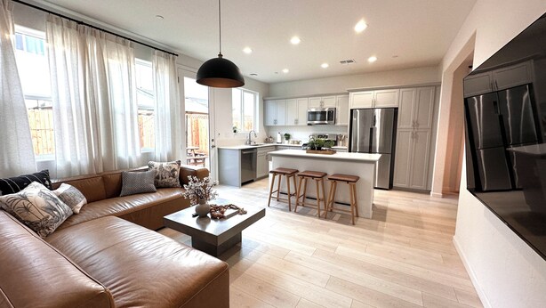 A modern kitchen with stainless steel appliances, a central island with stools, and a dining area with a brown leather sofa and a coffee table. - New! 4BR Vacation Home, Tub, BBQ, EV, close to Sequoia (Visalia)