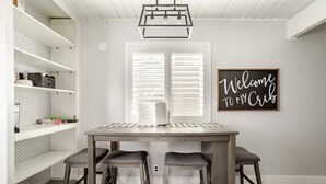 A dining area with a wooden table and two stools, a white shelving unit with storage, and a 'Welcome to My Crib' sign on the wall.