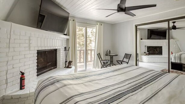 A bedroom with a white brick fireplace, a ceiling fan, a TV mounted above the fireplace, and a bed with a striped blanket.