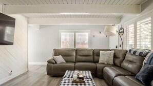 A living room with a brown leather sectional sofa, a checkered coffee table, and a large window with blinds.
