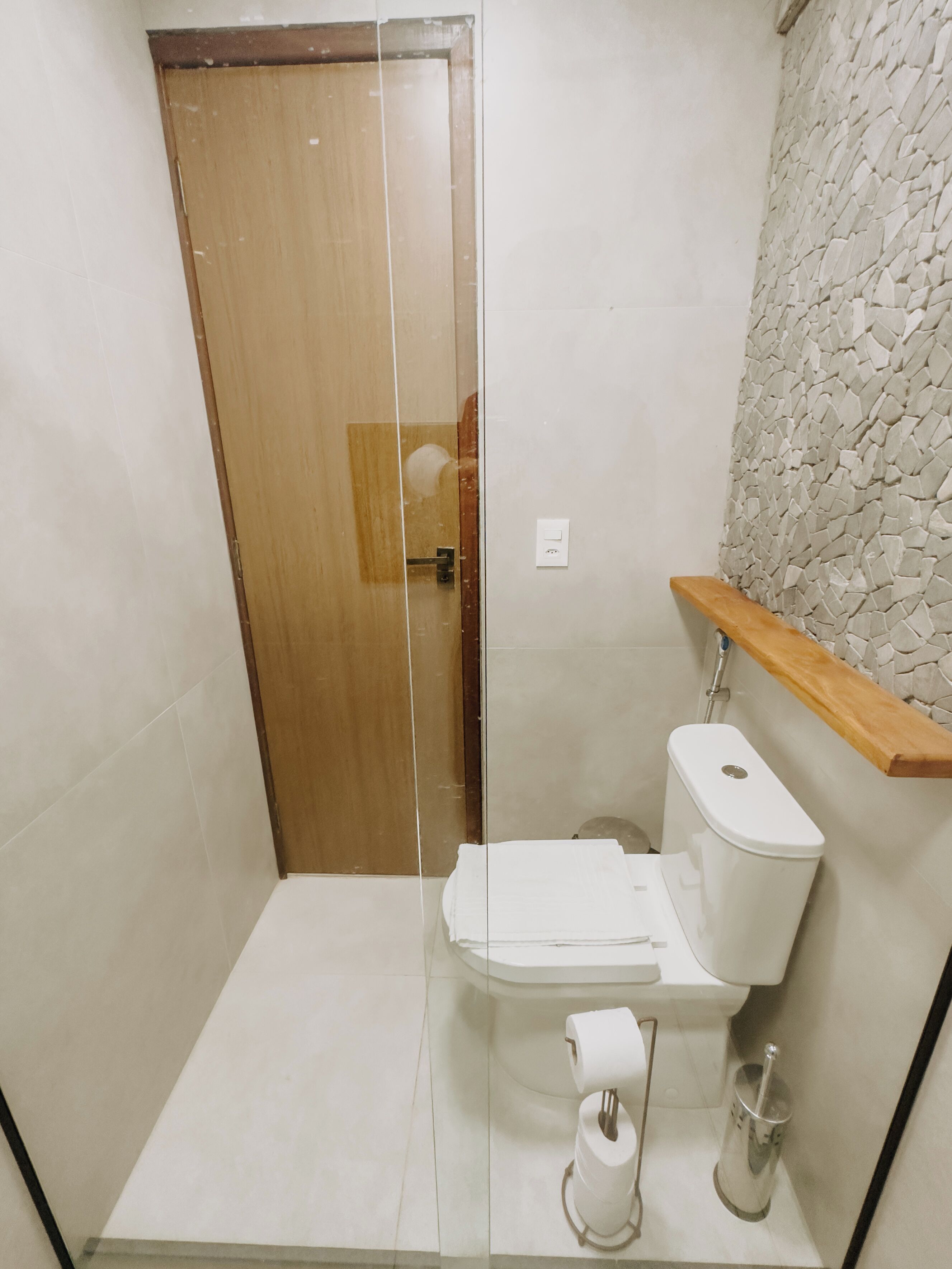 Ein Badezimmer mit Holztür, Toilette in Weiß, Duschkabine aus Glas und Wand mit Steinmuster.