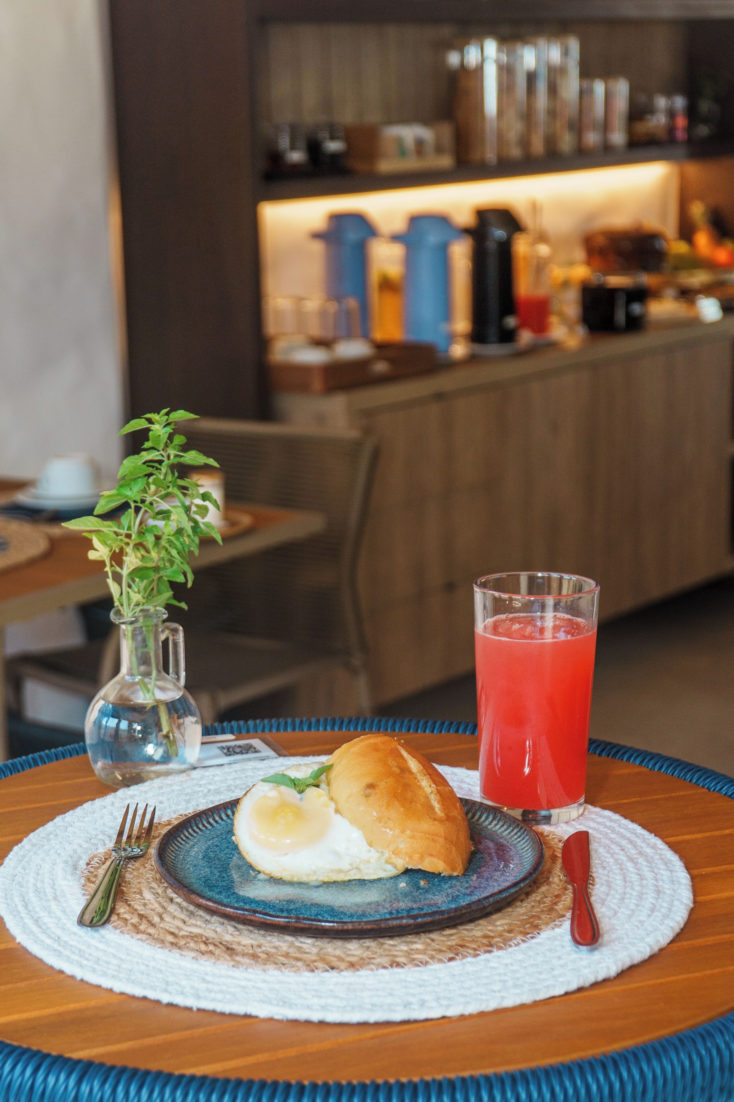 A breakfast setting with a plate of food, a glass of red juice, and a small plant on a woven placemat.