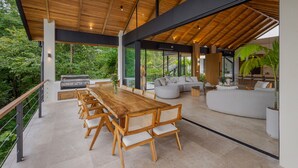 A spacious outdoor dining area with a wooden table and chairs, a built-in grill, and a large window overlooking a lush garden. - Villa Bahia (Cóbano)
