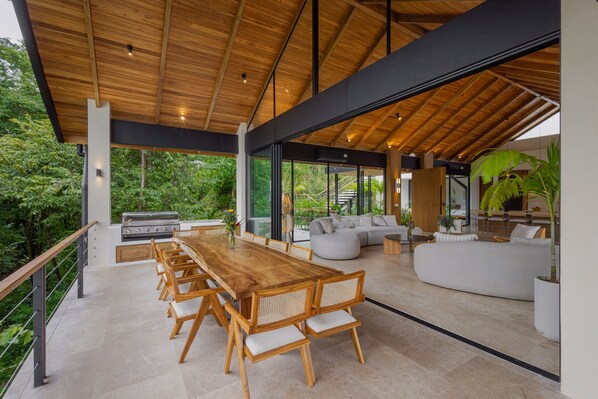 A spacious outdoor dining area with a wooden table and chairs, a built-in grill, and a large window overlooking a lush garden.