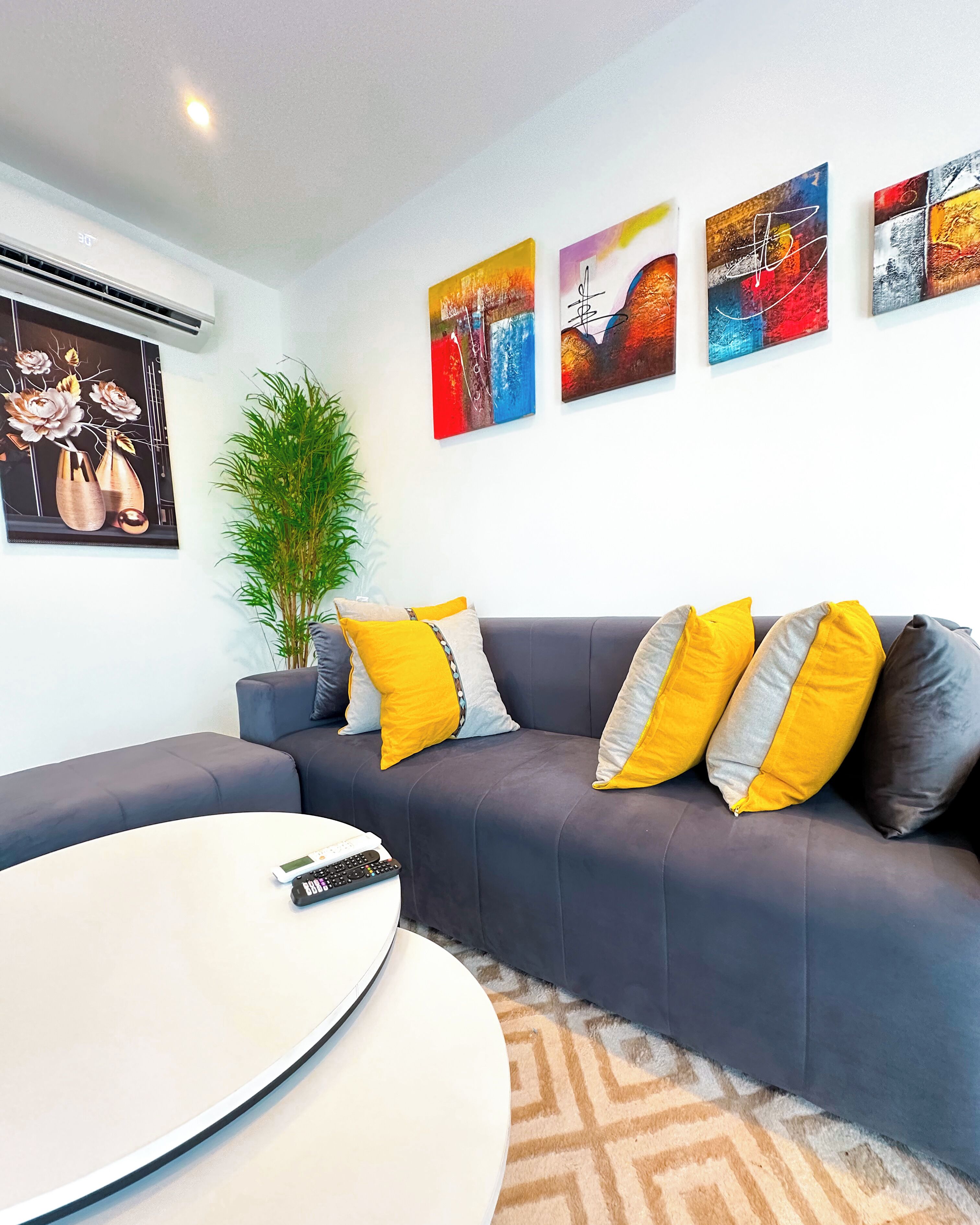 A modern living room with a grey sofa, yellow and grey cushions, a white coffee table, and a remote control. There are four framed artworks on the wall.
