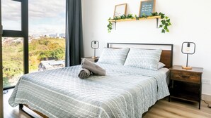 A neatly made bed with a quilted bedspread and pillows, a bedside table with a lamp, and a view of the city through large windows.