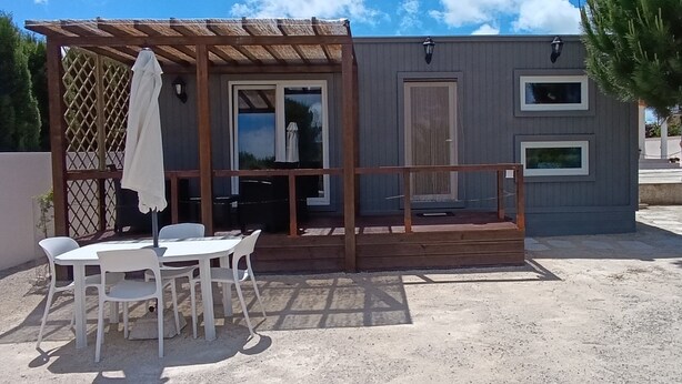 A small, single-story building with a covered patio area, a table with chairs, and a wooden deck.