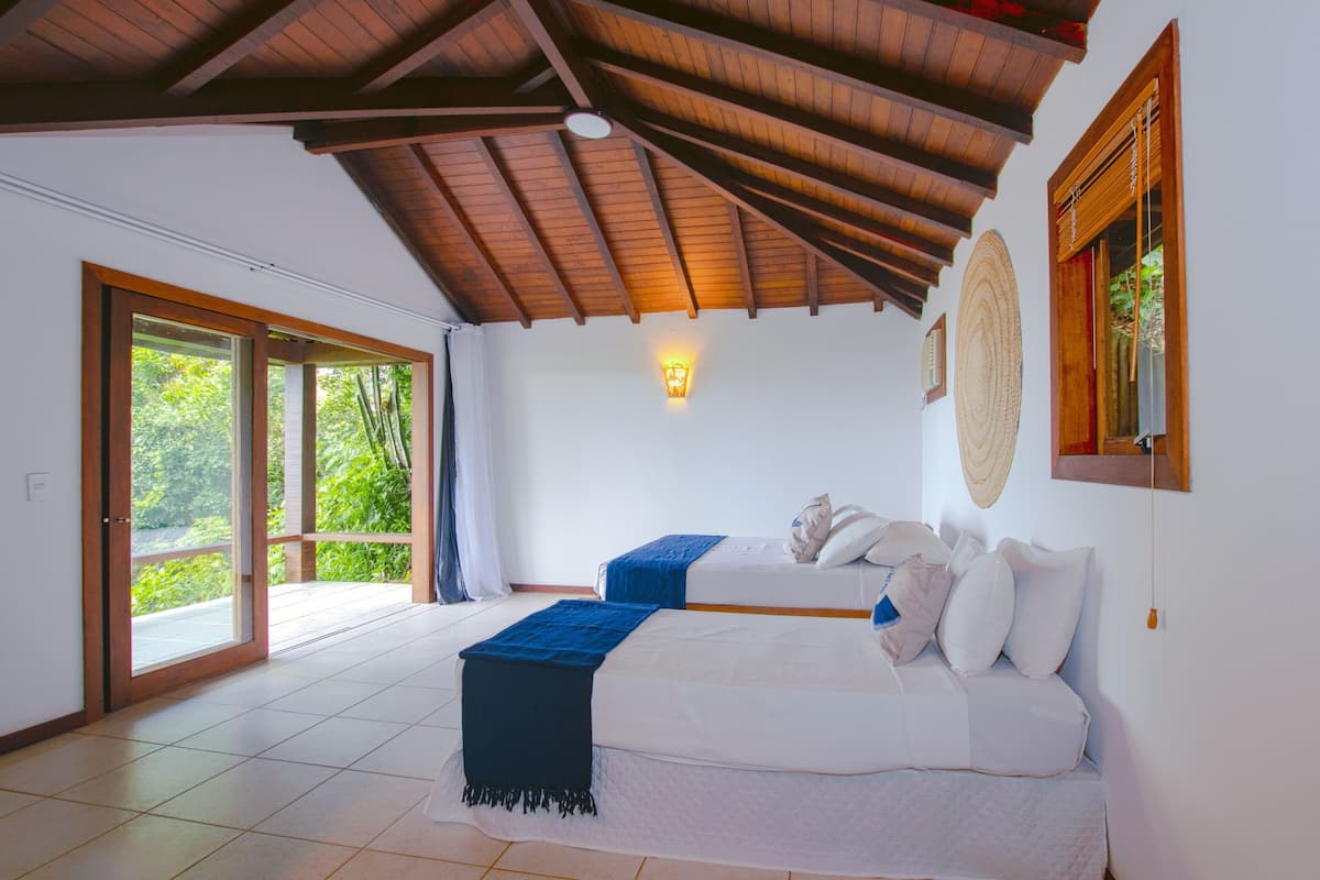 Una habitación amplia con techo alto, una cama grande, una cama secundaria más pequeña y vistas a través de un ventanal corredero.