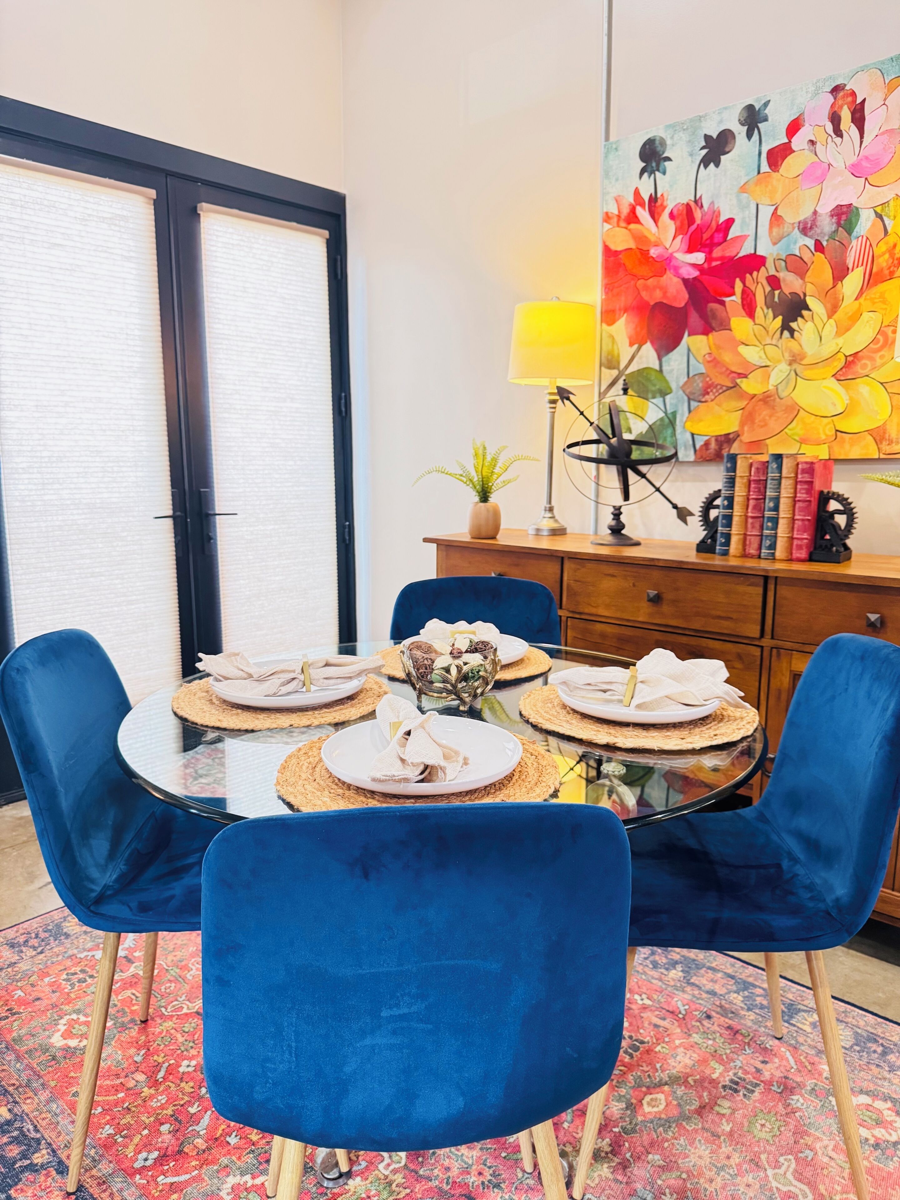 Un coin repas avec une table ronde en verre, des chaises en velours bleu, un tableau floral aux couleurs vives, une lampe et une bibliothèque.