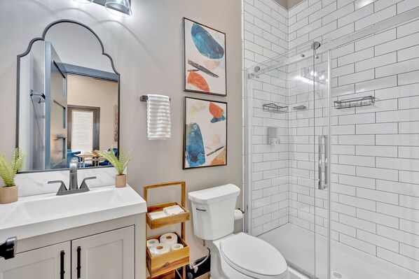 Une salle de bain moderne avec des toilettes blanches, une douche en verre, un lavabo avec un miroir et deux tableaux abstraits encadrés.