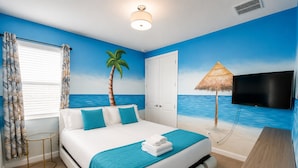 Un dormitorio con cama, televisor, ventana con cortinas, puerta y un mural en la pared con temática de playa.