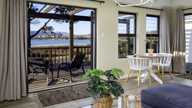 A room with a view of the sea, a wooden deck, and a dining area with white chairs and a table. - Fisherhaven - Four Bedroom Apartment, Sleeps 8 (Hermanus)