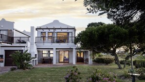 A two-story house with a balcony, a covered patio, and a well-maintained garden with purple flowers. - Fisherhaven - Four Bedroom Apartment, Sleeps 8 (Hermanus)