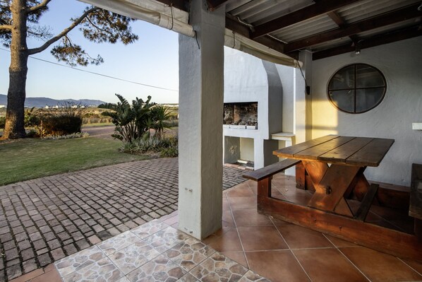 An outdoor patio area with a wooden table and benches, surrounded by a paved walkway and a grassy lawn.