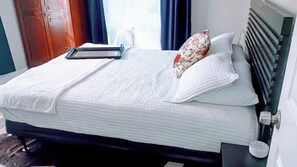 Ein ordentlich bezogenes Bett mit weißer Bettwäsche, einem Blumenkissen und einem Nachttisch mit einem weißen Gegenstand darauf.