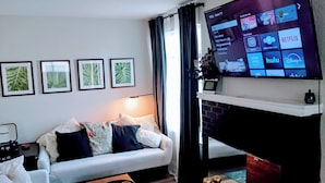 A living room with a flat-screen TV mounted on the wall, a white sofa with floral cushions, and framed botanical prints on the wall. - 2BR/1BA near Jax Zoo -- Welcome, Train Enthusiasts!!  (Jacksonville)