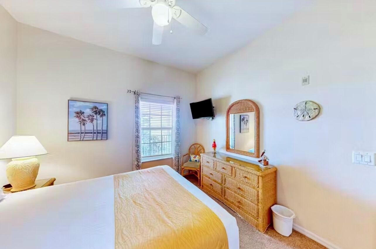 Una camera d'albergo con un letto, una cassettiera, una televisione, una finestra con tende e una ventola a soffitto.