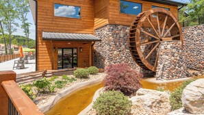 A wooden building with a large water wheel, a stone wall, and a small pond with bushes.