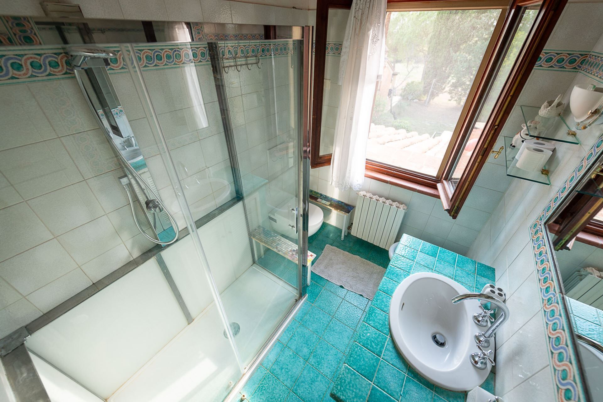 Un baño con ducha acristalada, lavamanos con espejo y ventana que proporciona luz natural.