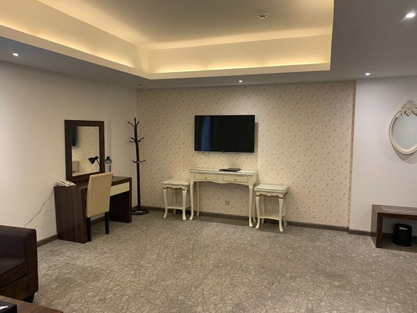 Una habitación de hotel moderna con una televisión de pantalla plana grande montada en la pared, un escritorio con una silla, un perchero y una mesita de noche con una lámpara.