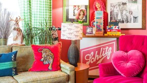 A vibrant living room with a red sofa, a green and white checkered curtain, a decorative rabbit figurine, a tiger-themed pillow, a lamp, and a shelf with books and a PlayStation. - Betty Blue House (Reno)