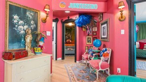 A vibrant room with a pink wall, a framed painting, a dresser with a red suitcase, and a blue chair. - Betty Blue House (Reno)