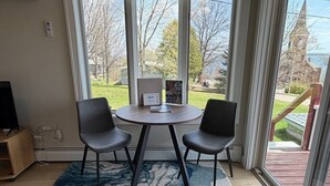Een kamer met een ronde tafel, twee stoelen, een vloerkleed en uitzicht op een gebouw en bomen door grote ramen.
