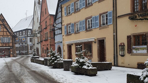 Une rue enneigée bordée d’immeubles aux façades typiques de l’architecture européenne, notamment des maisons à colombages, et d’un restaurant doté d’une terrasse.
