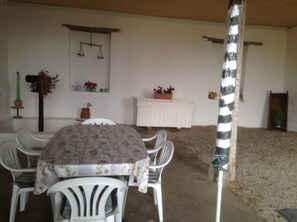 Un coin repas avec une table recouverte d’une nappe à motifs floraux, entourée de chaises blanches. On y trouve un buffet blanc sur lequel sont posées des plantes en pot, et une balance accrochée au mur.