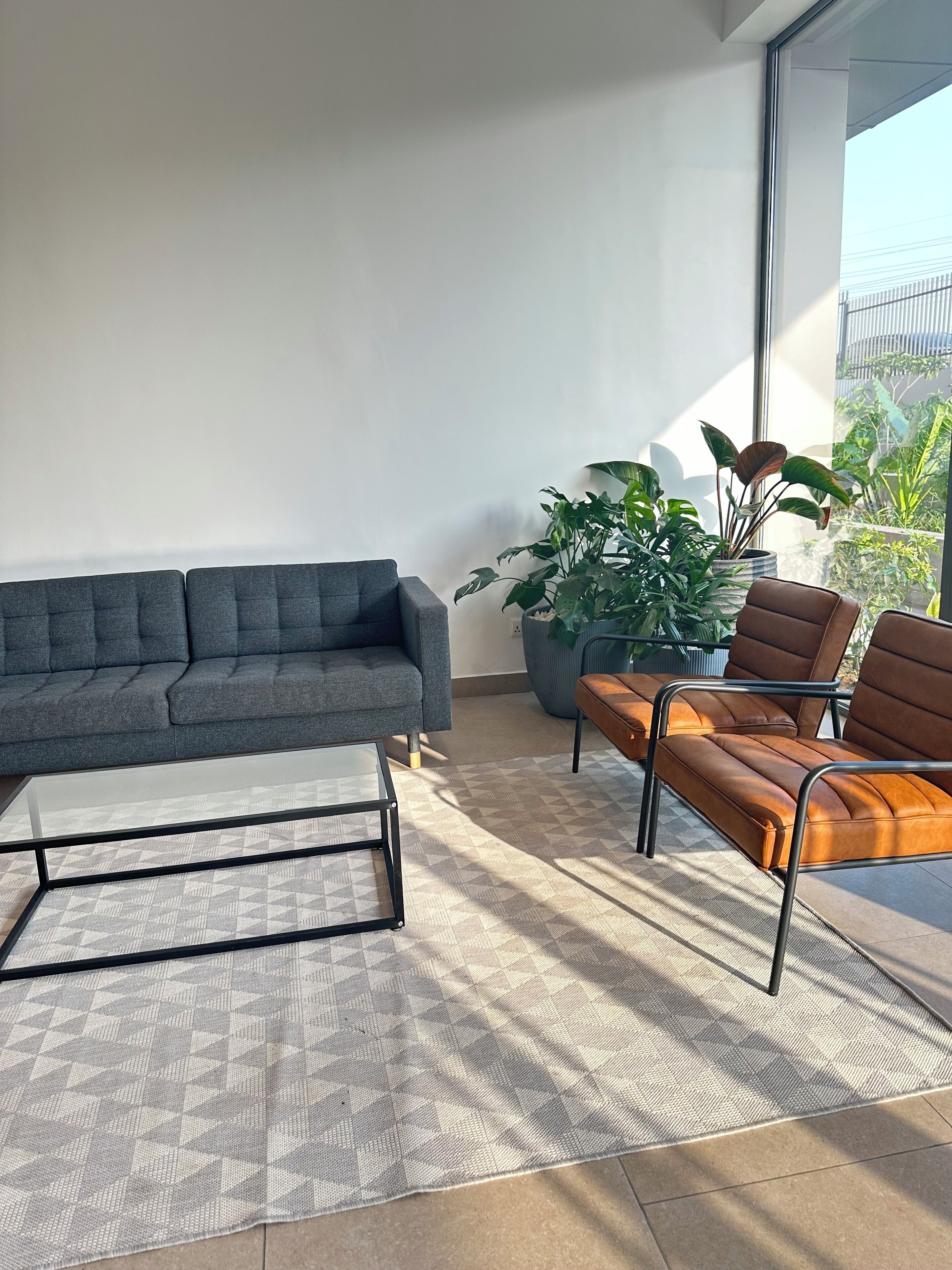 Un salon moderne avec un canapé gris, une table basse et un canapé en cuir marron. On y trouve une grande plante en pot et une vue sur la verdure à l’extérieur.