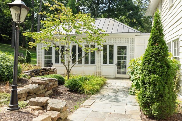 A conservatory with a stone pathway leading to a white door, surrounded by greenery and a lamp post.