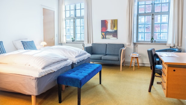 Une chambre d’hôtel avec un lit, un banc bleu, un canapé, un bureau et deux fenêtres avec des rideaux.
