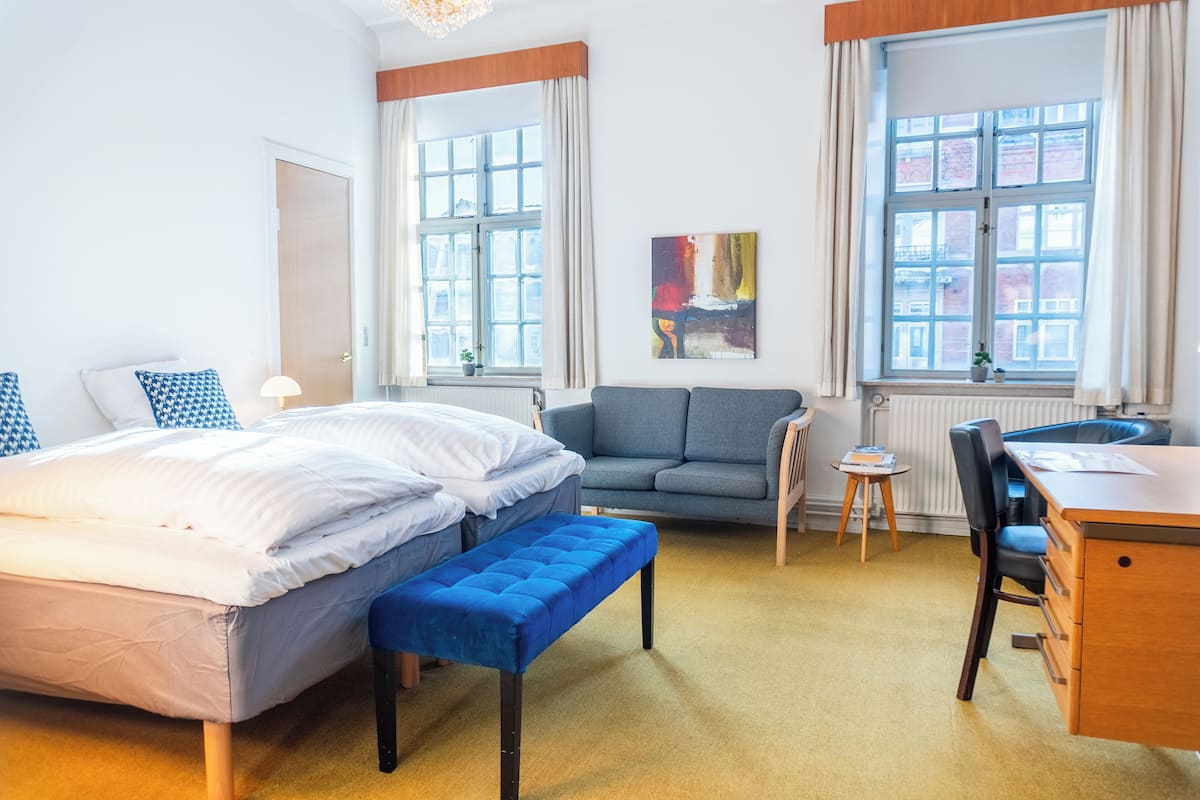 Une chambre d’hôtel avec un lit, un banc bleu, un canapé, un bureau et deux fenêtres avec des rideaux.