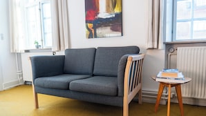Un salon moderne avec un canapé gris, une petite table d’appoint en bois sur laquelle sont posés des livres, un tableau abstrait aux couleurs vives accroché au mur, et une fenêtre avec des rideaux.