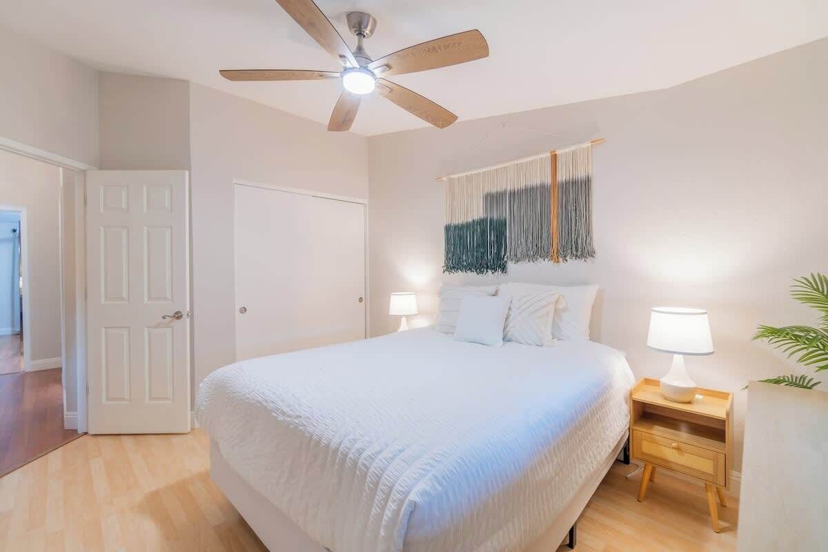 A bedroom with a bed, a nightstand, a wall-mounted lamp, a ceiling fan, and a door.