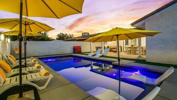 An outdoor swimming pool with lounge chairs and umbrellas, surrounded by a white wall and a building with a patio. - Ultimate Coachella Stay: Pool & Fun & pickleball (Coachella)