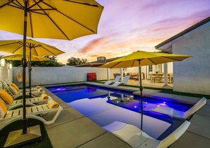 An outdoor swimming pool with lounge chairs and umbrellas, surrounded by a white wall and a building with a patio.