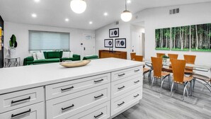 A modern kitchen with a white island, green sofa, and dining area with wooden chairs. - Ultimate Coachella Stay: Pool & Fun & pickleball (Coachella)