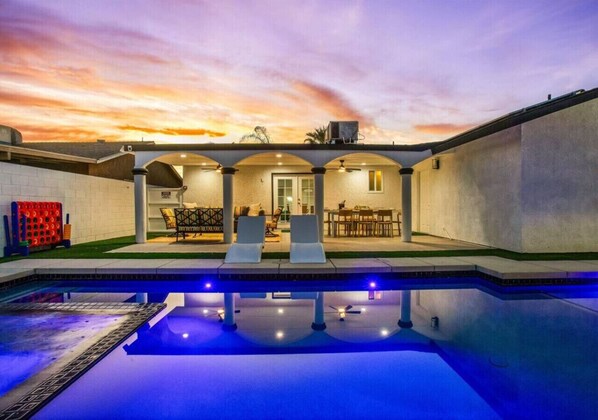 A swimming pool with a sunset view, a covered patio with seating, and a building with a flat roof.