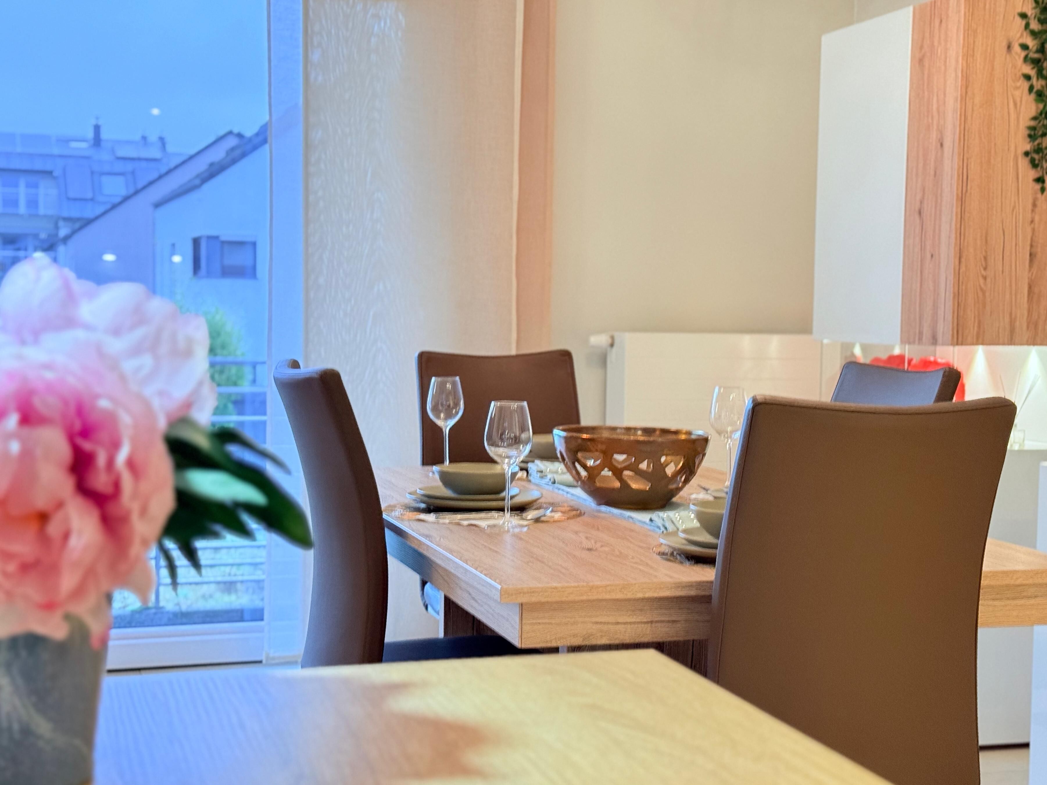 A dining area with a wooden table, four chairs, a vase with a flower, and a view of buildings outside.