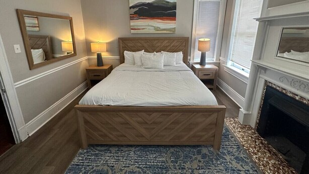 A neatly arranged bedroom with a large bed, bedside tables, a fireplace, and a painting on the wall.