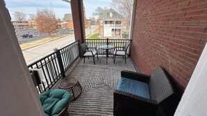 A balcony with wicker furniture, a patterned rug, and a view of a street and buildings.