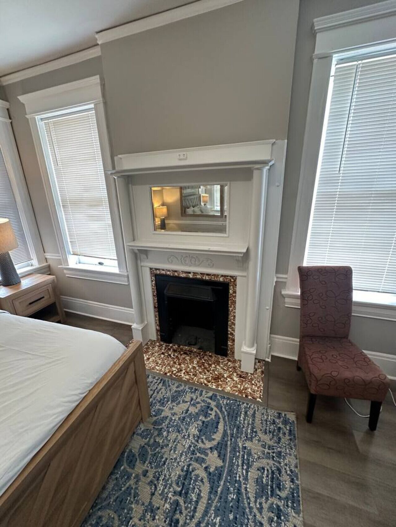 A bedroom with a bed, a fireplace, a chair, and a window with blinds.
