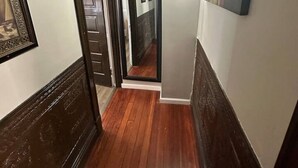 A narrow hallway with wooden flooring, a door on the left, and a wall-mounted light fixture on the right.