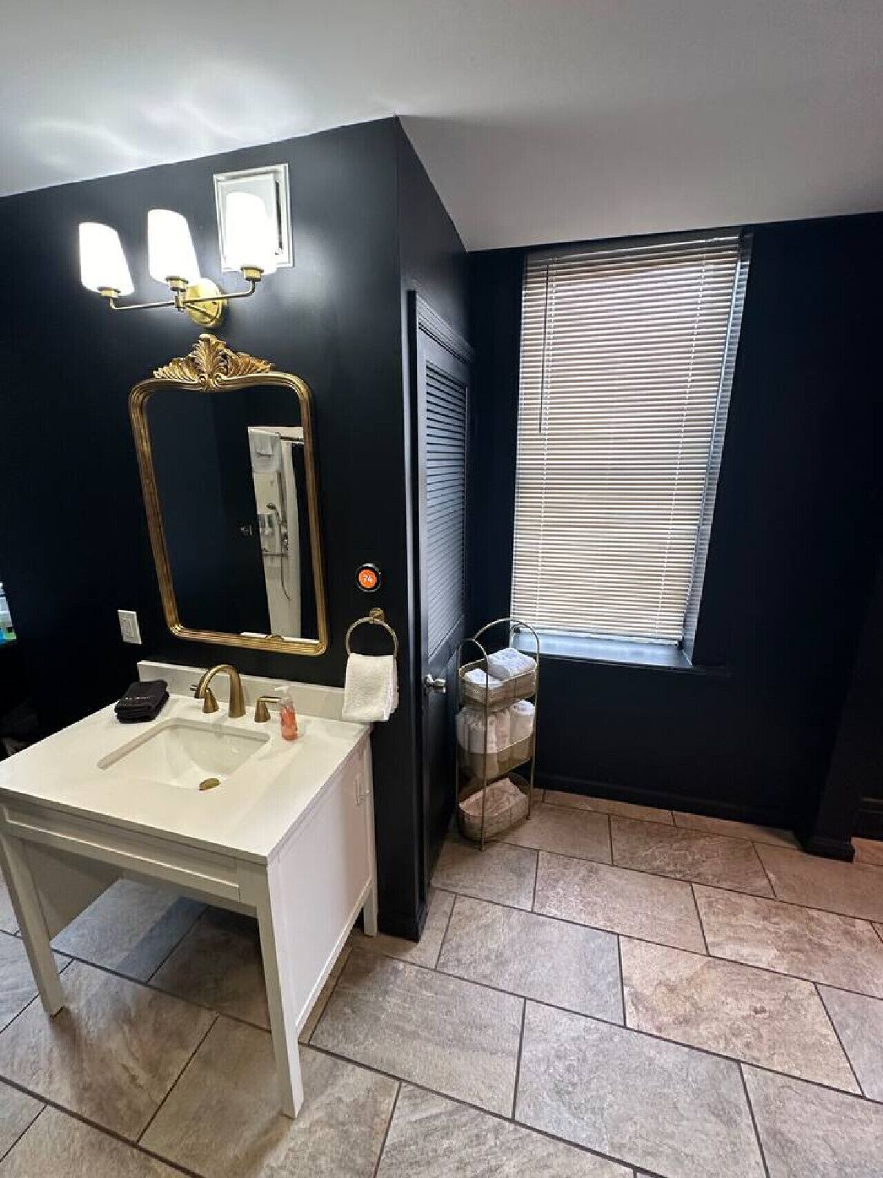 A bathroom with a white sink, gold faucets, a large mirror with a gold frame, and a window with blinds.