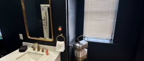A bathroom with a white sink, gold faucets, a large mirror with a gold frame, and a window with blinds.
