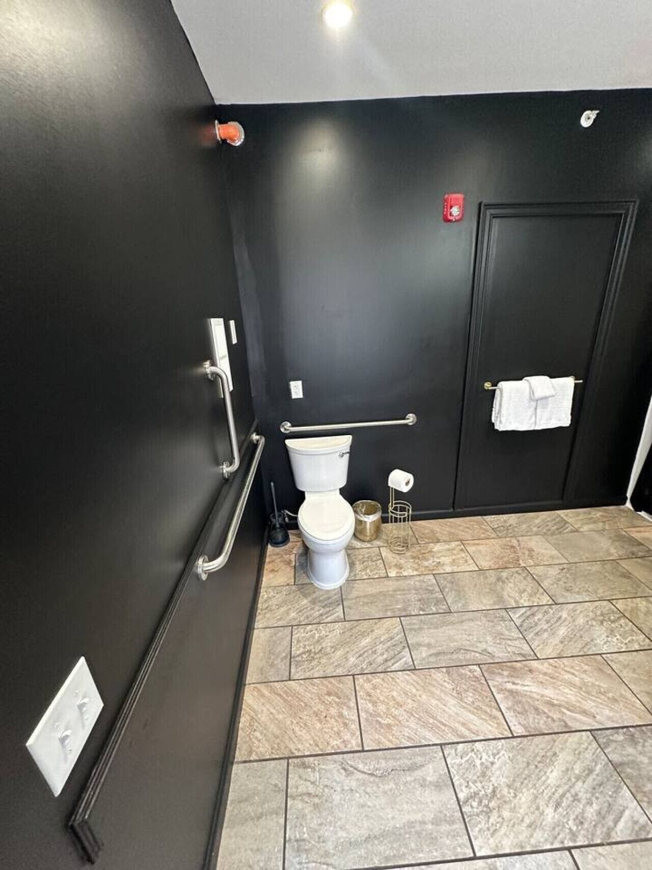Un baño con inodoro, barra de apoyo, toallero y suelo de baldosas.