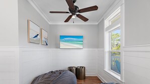 A bedroom with a ceiling fan, a large window with a view of the outdoors, a bed with a gray blanket, and two cylindrical objects on the floor.