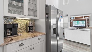 A modern kitchen with a stainless steel refrigerator, a coffee maker, and a microwave. There is a built-in oven and a microwave above the counter. The kitchen has white cabinets and a herringbone patterned backsplash.